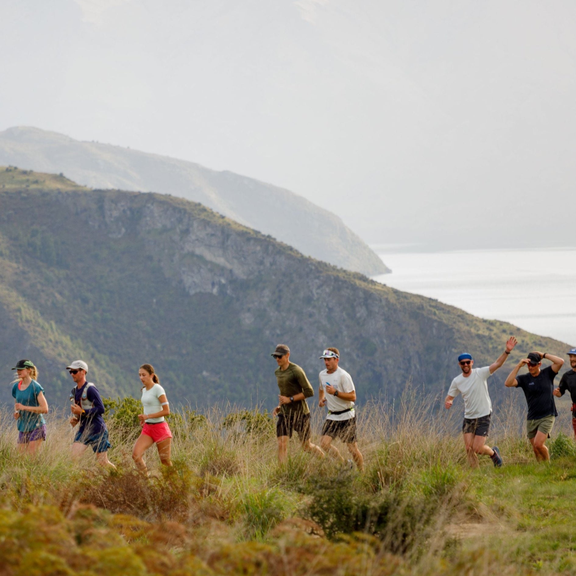 A group of runners participates in a trail race on a grassy mountainside with a dramatic coastal mountain backdrop.