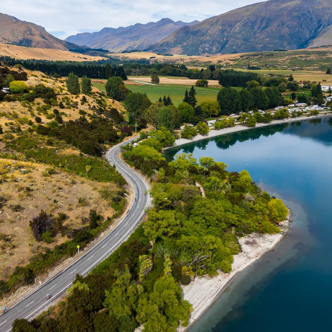 Aerial shot of a road winding along a turquoise lake with mountains in the background.