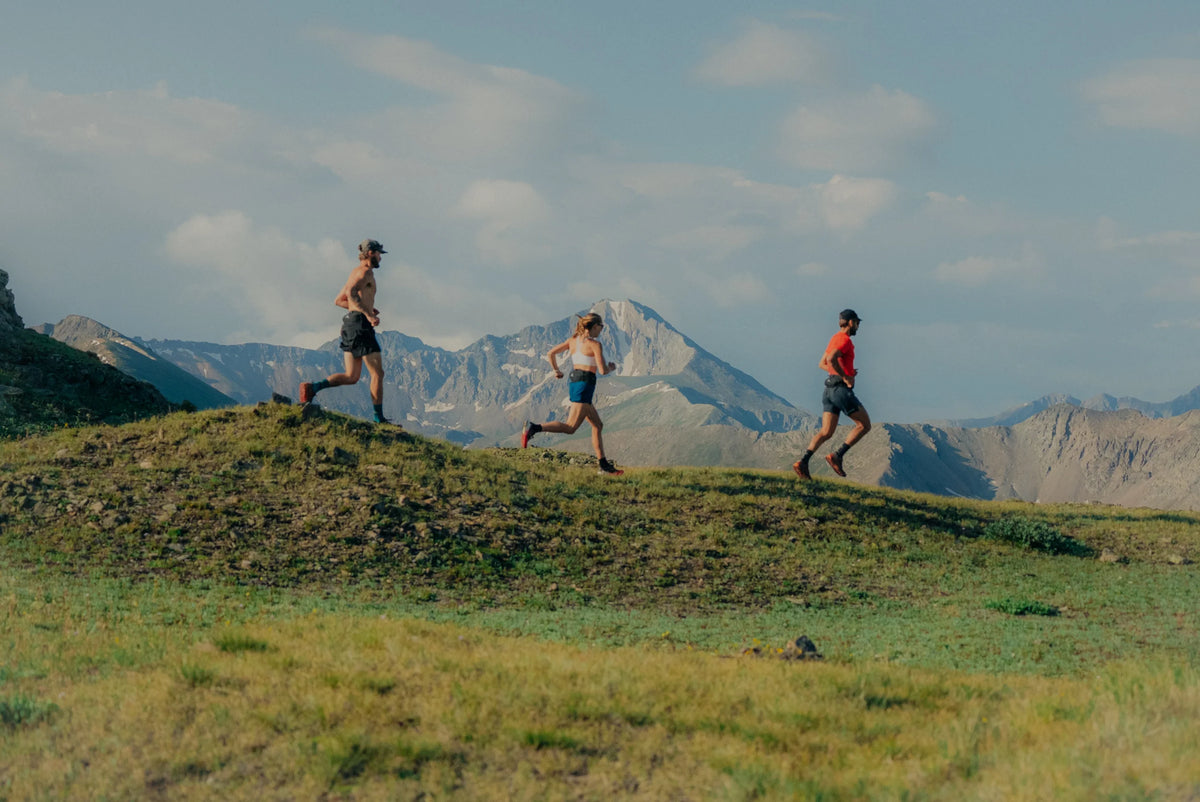 Three trail runners on a grassy ridge with mountains behind
