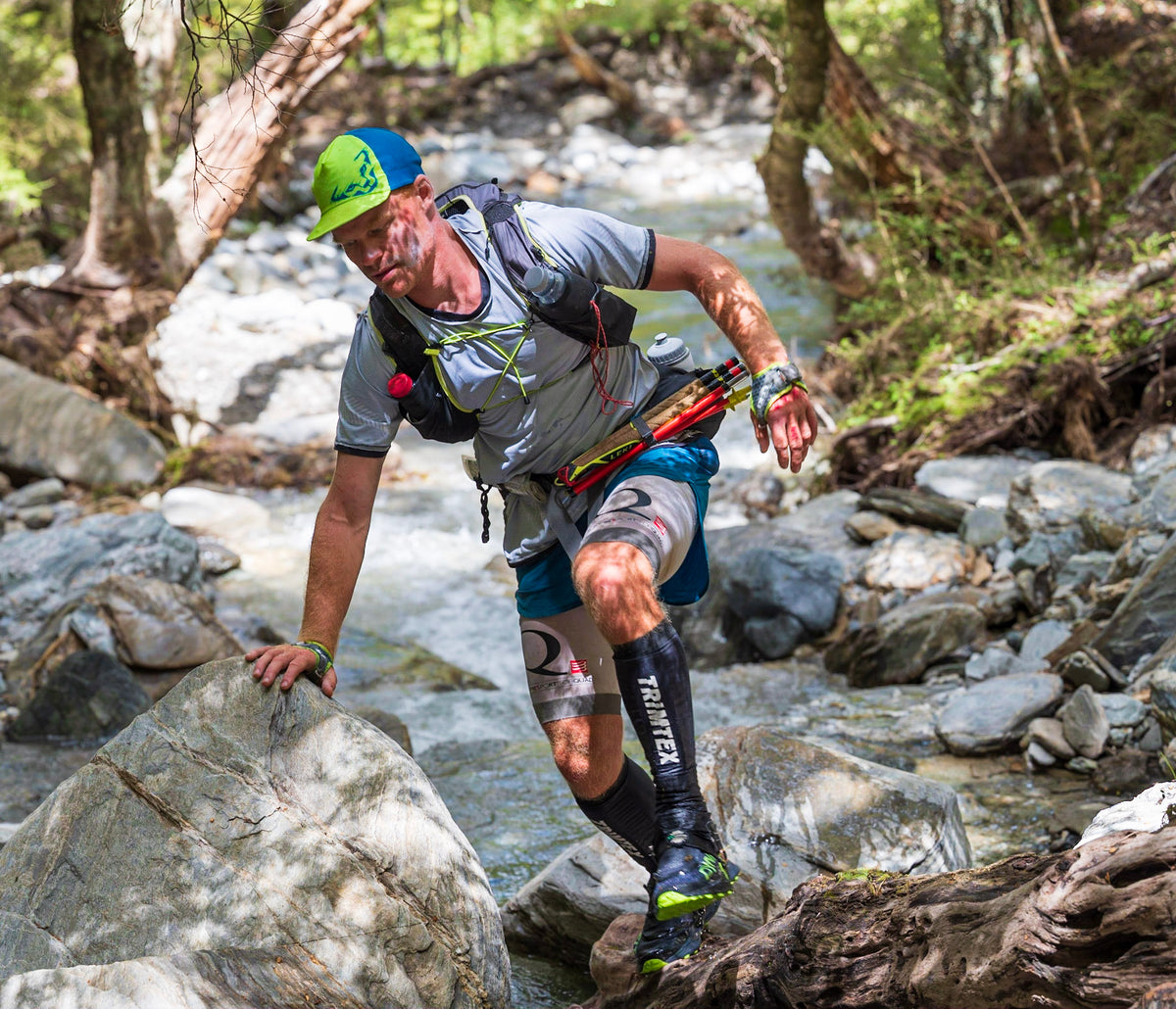A male trail runner navigating a rocky stream in a forest, wearing a blue and green cap, gray shirt, blue shorts, and black compression sleeves.