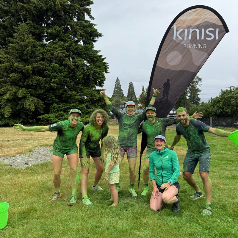 The Kinisi Running crew covered in green powder after the Sport Otago Wānaka Colour Run, in front of the Kinisi Running flag
