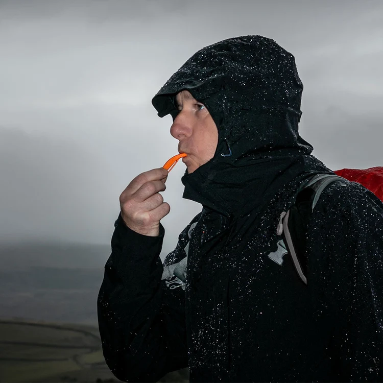 A person in a black hooded rain jacket standing outdoors in the rain, holding an orange object near their mouth. A red backpack is partially visible behind them, with a cloudy gray sky in the background.
