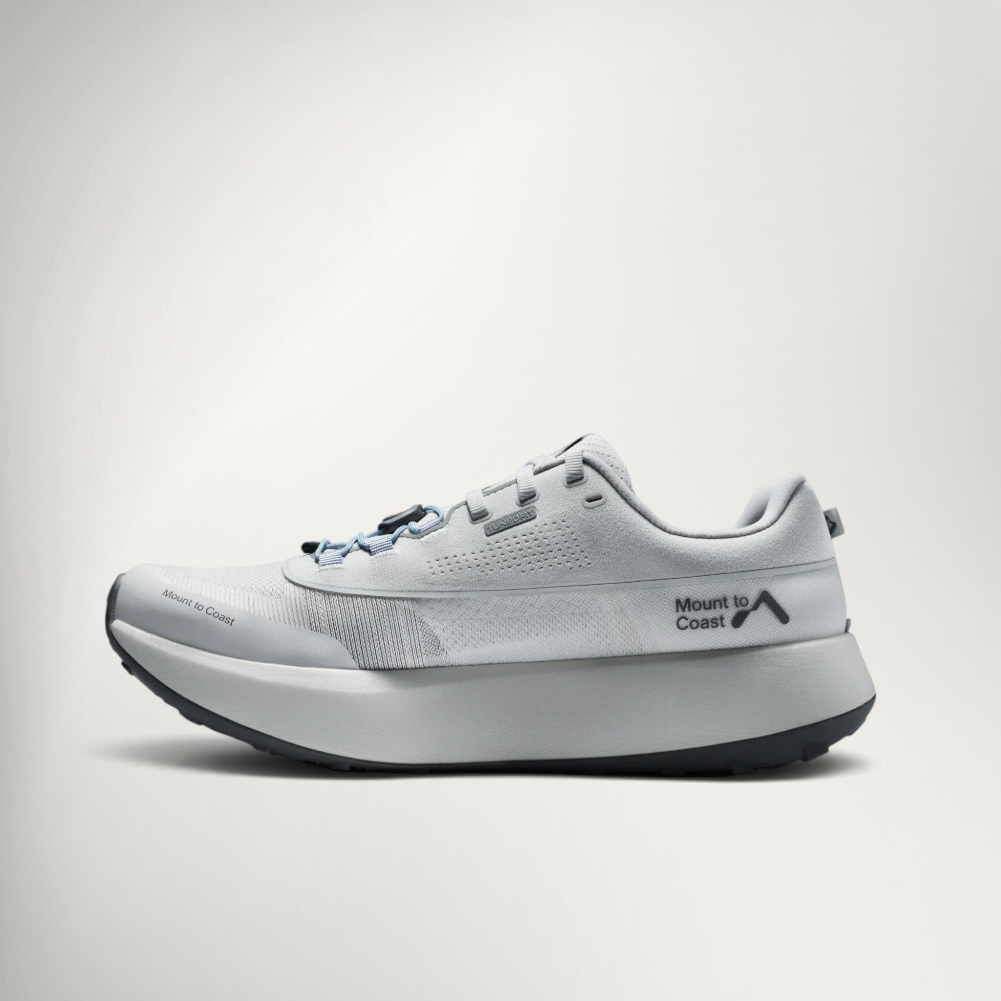 A light gray and white Mount To Coast running shoe with a thick white midsole and a black logo on the side, shown against a light gray background.
