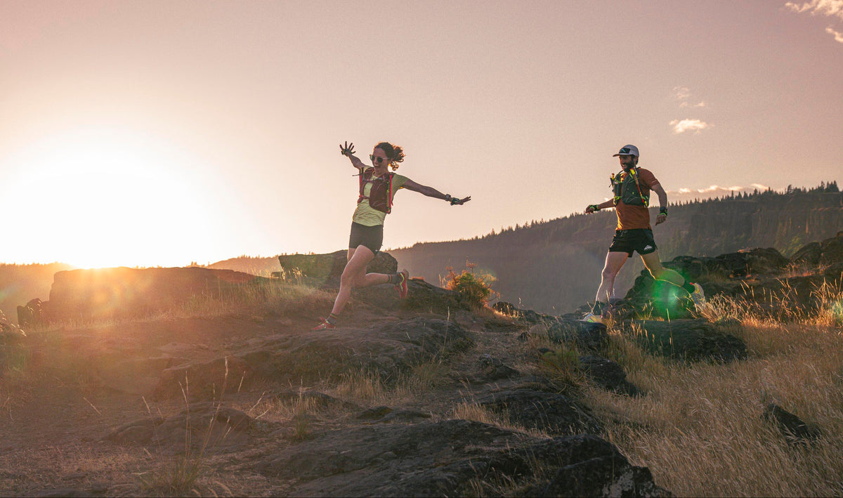 Two trail runners wearing Osprey running vests leaping across a hilltop at sunset
