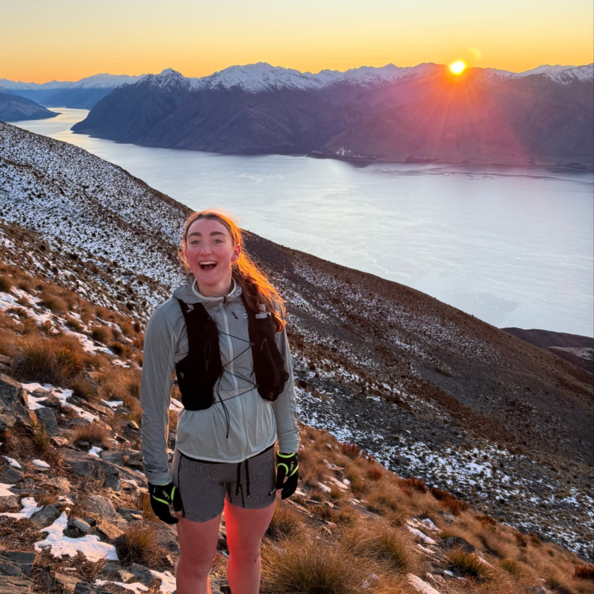 Piper, trail running on a snowy mountain with a lake behind and the sun rising over the mountain in the background.