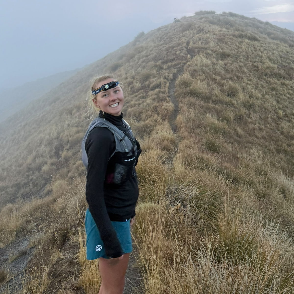 Scarlett, trail running up a grassy ridge with the mountains slightly shrouded in fog.