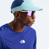 A man in profile wearing a light blue and navy blue The North Face cap, white-framed sunglasses with pink mirrored lenses, and a royal blue The North Face t-shirt against a clean white background.