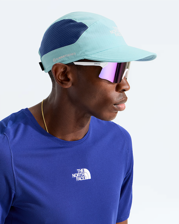 A man in profile wearing a light blue and navy blue The North Face cap, white-framed sunglasses with pink mirrored lenses, and a royal blue The North Face t-shirt against a clean white background.