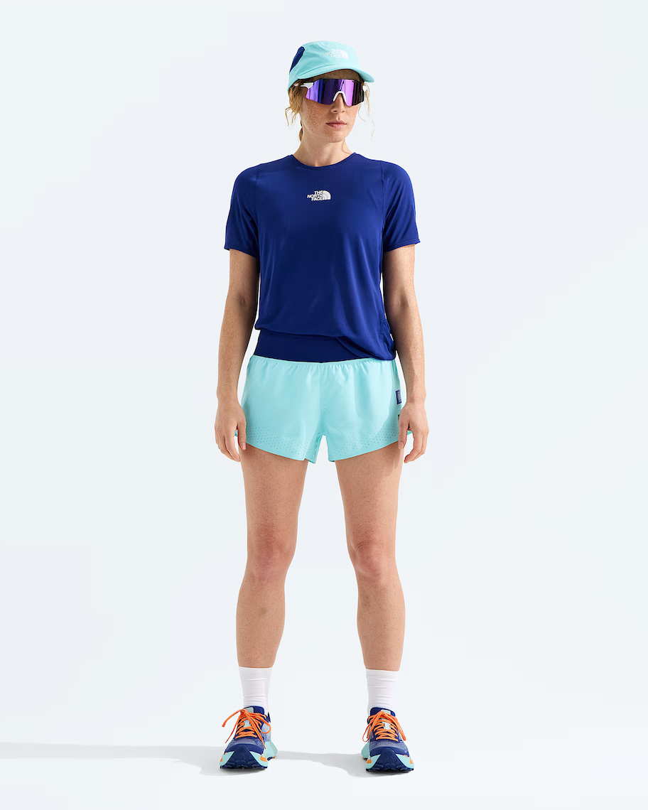 A model stands against a light gray studio background wearing a navy blue short-sleeve athletic t-shirt with a small white chest logo, light aqua running shorts, a matching light aqua visor, reflective sunglasses, white socks, and blue running shoes with orange laces. The t-shirt brand is The North Face.