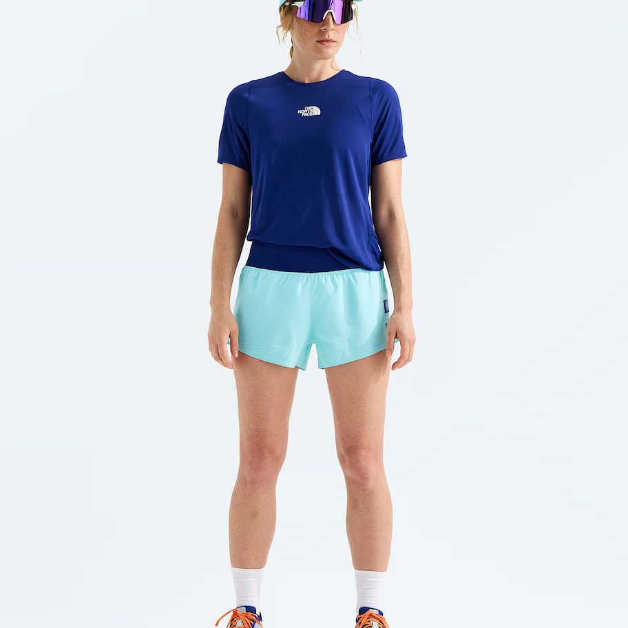A model stands against a light gray studio background wearing a navy blue short-sleeve athletic t-shirt with a small white chest logo, light aqua running shorts, a matching light aqua visor, reflective sunglasses, white socks, and blue running shoes with orange laces. The t-shirt brand is The North Face.
