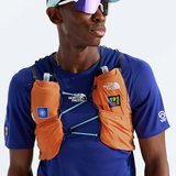 A male athlete wearing a blue The North Face performance shirt and a light blue cap, wearing an orange hydration vest with two water bottles in the side pockets. The background is a clean white studio backdrop.