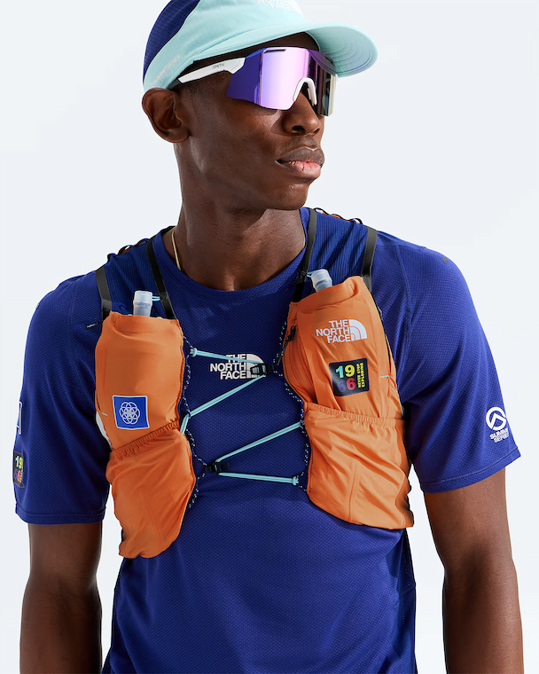 A male athlete wearing a blue The North Face performance shirt and a light blue cap, wearing an orange hydration vest with two water bottles in the side pockets. The background is a clean white studio backdrop.