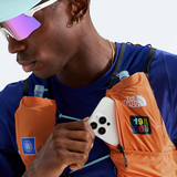 A close-up lifestyle shot of a man wearing an orange The North Face hydration pack over a blue athletic shirt, holding a white smartphone in front of the pack pocket. The man is also wearing a light blue cap and mirrored sunglasses.