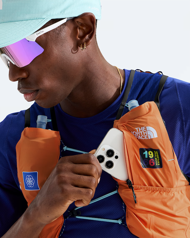A close-up lifestyle shot of a man wearing an orange The North Face hydration pack over a blue athletic shirt, holding a white smartphone in front of the pack pocket. The man is also wearing a light blue cap and mirrored sunglasses.
