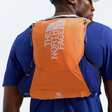 A man wearing a blue athletic shirt and a light blue visor, shown from behind with a bright orange The North Face hydration pack on his back. The pack features the brand's logo in white on the upper back and a small white logo near the bottom.