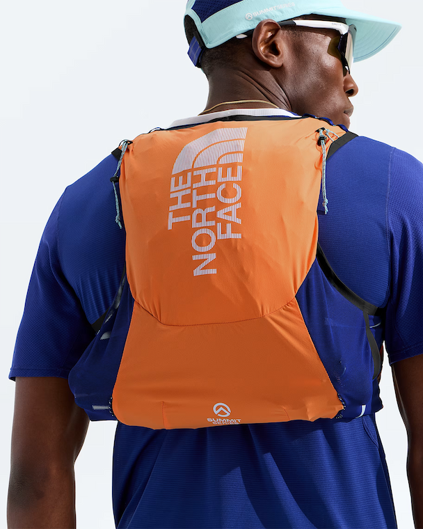 A man wearing a blue athletic shirt and a light blue visor, shown from behind with a bright orange The North Face hydration pack on his back. The pack features the brand's logo in white on the upper back and a small white logo near the bottom.