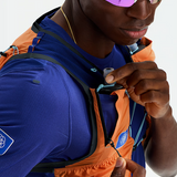 Close-up of a runner wearing a blue and orange hydration vest over a blue athletic shirt, with a small white hydration reservoir attached at the chest.