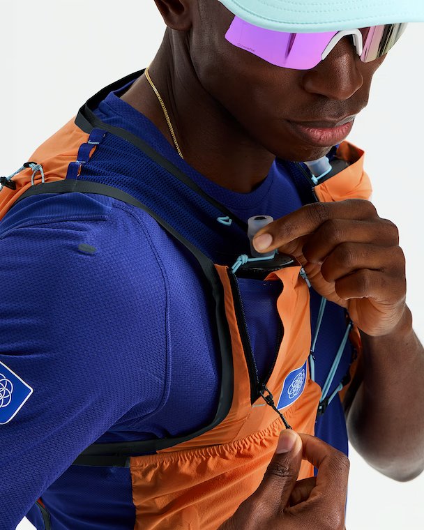 Close-up of a runner wearing a blue and orange hydration vest over a blue athletic shirt, with a small white hydration reservoir attached at the chest.