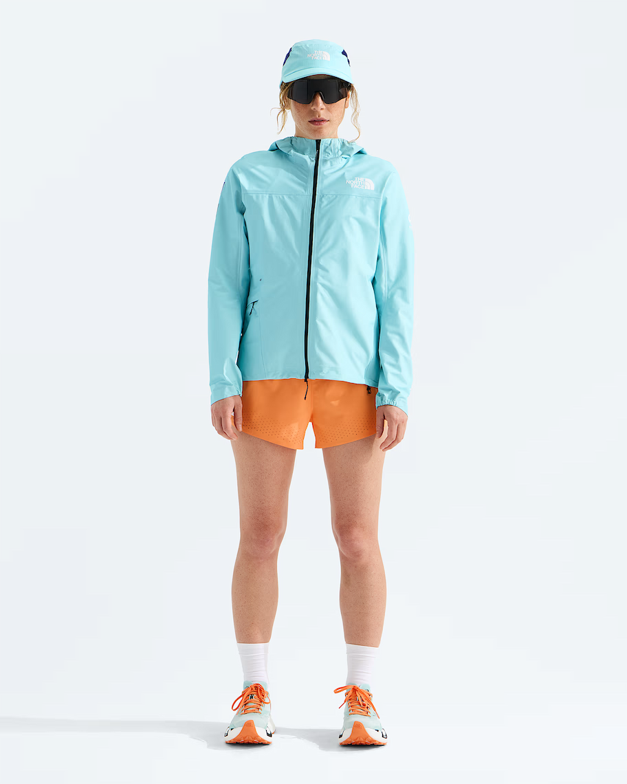 A model stands against a white studio background wearing a light blue hooded jacket, orange shorts, white socks, and orange and light blue running shoes, with a matching light blue cap and dark sunglasses.