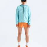 A model stands against a white studio background wearing a light blue hooded jacket, orange shorts, white socks, and orange and light blue running shoes, with a matching light blue cap and dark sunglasses.