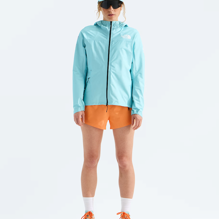 A model stands against a white studio background wearing a light blue hooded jacket, orange shorts, white socks, and orange and light blue running shoes, with a matching light blue cap and dark sunglasses.