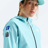 A woman wearing a light blue hooded jacket and matching visor, photographed against a clean white studio background. The jacket has visible patches on the sleeve and a small chest logo, and the visor shows The North Face branding.