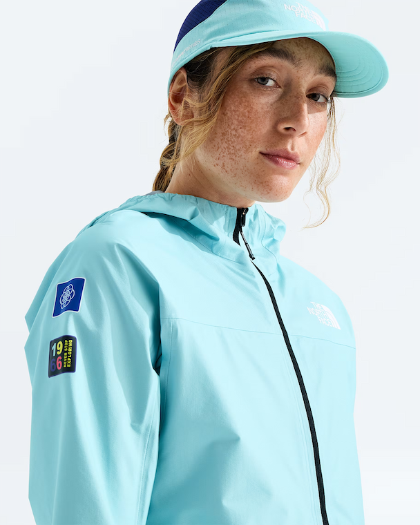 A woman wearing a light blue hooded jacket and matching visor, photographed against a clean white studio background. The jacket has visible patches on the sleeve and a small chest logo, and the visor shows The North Face branding.