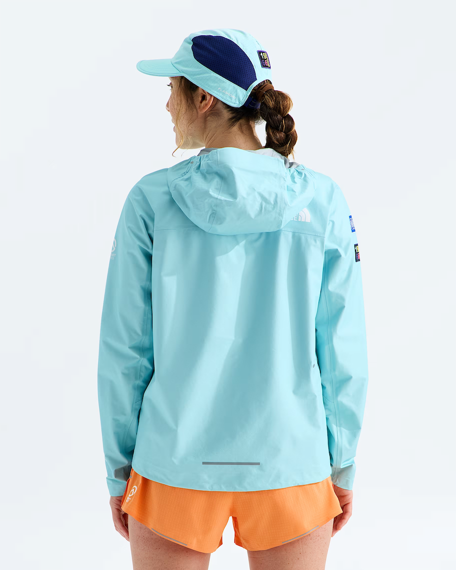 A woman shown from the back wearing a light blue hooded jacket, orange shorts, and a navy blue and light blue cap, photographed against a clean white studio background.