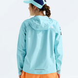 A woman shown from the back wearing a light blue hooded jacket, orange shorts, and a navy blue and light blue cap, photographed against a clean white studio background.