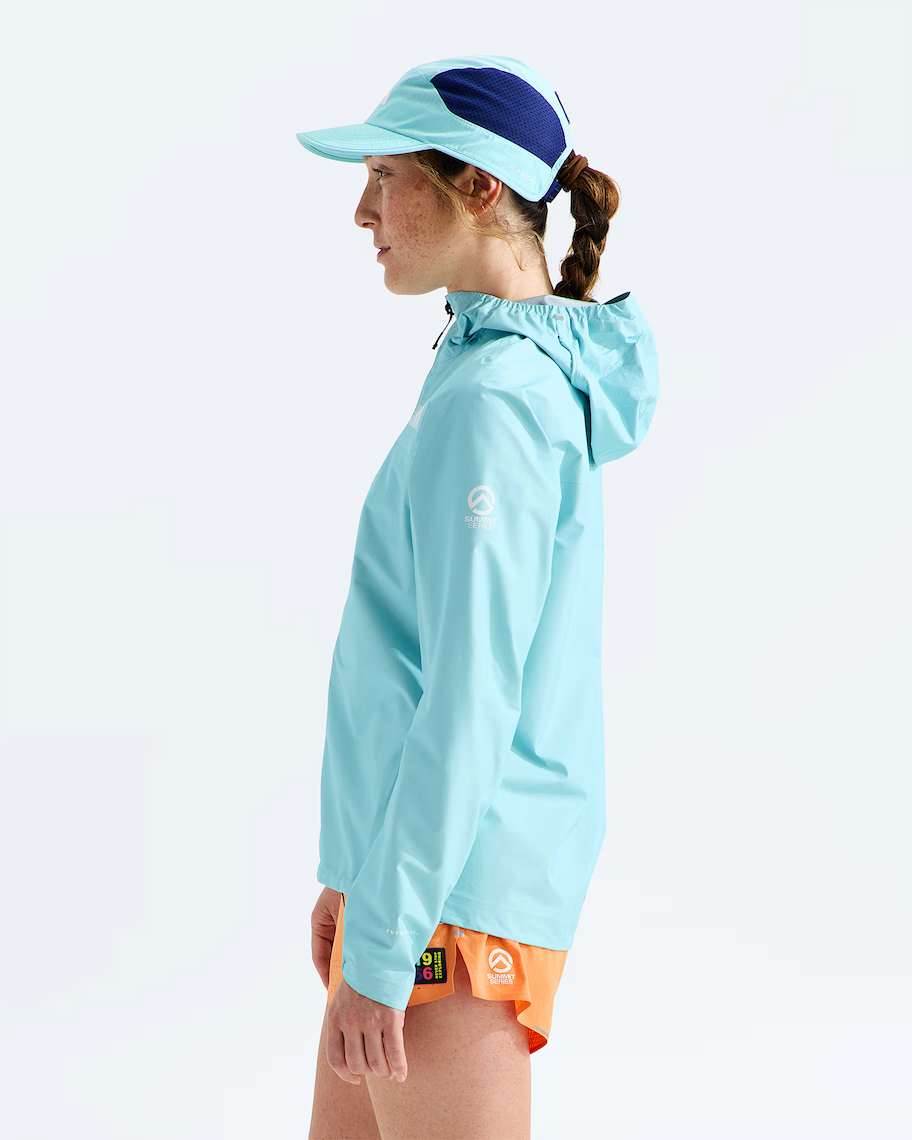 A woman in profile wearing a light blue hooded athletic jacket, a matching light blue visor with a dark blue top panel, and bright orange running shorts against a clean white studio background.