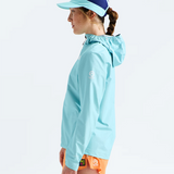 A woman in profile wearing a light blue hooded athletic jacket, a matching light blue visor with a dark blue top panel, and bright orange running shorts against a clean white studio background.