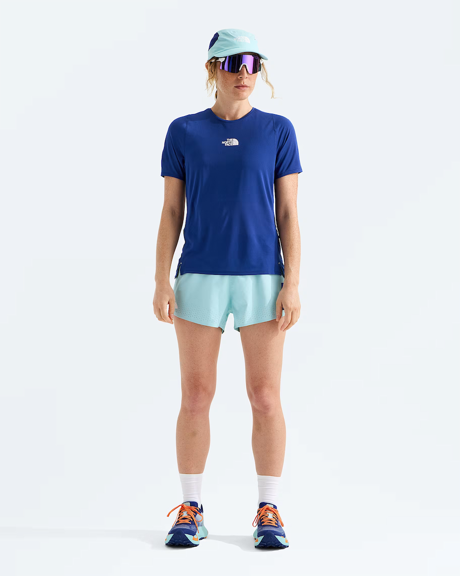 A model stands against a clean white studio background wearing a royal blue short-sleeve athletic T-shirt with a small white The North Face logo on the chest, light aqua running shorts, a pale blue cap, reflective sunglasses, white socks, and blue running shoes.