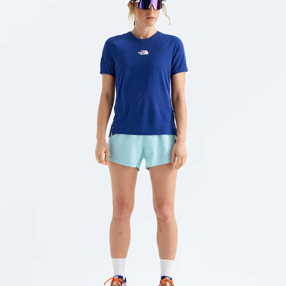 A model stands against a clean white studio background wearing a royal blue short-sleeve athletic T-shirt with a small white The North Face logo on the chest, light aqua running shorts, a pale blue cap, reflective sunglasses, white socks, and blue running shoes.