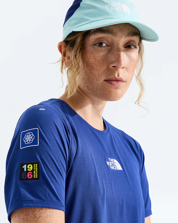 A close-up of a woman wearing a blue The North Face athletic t-shirt and a light blue cap, photographed against a clean white studio background.