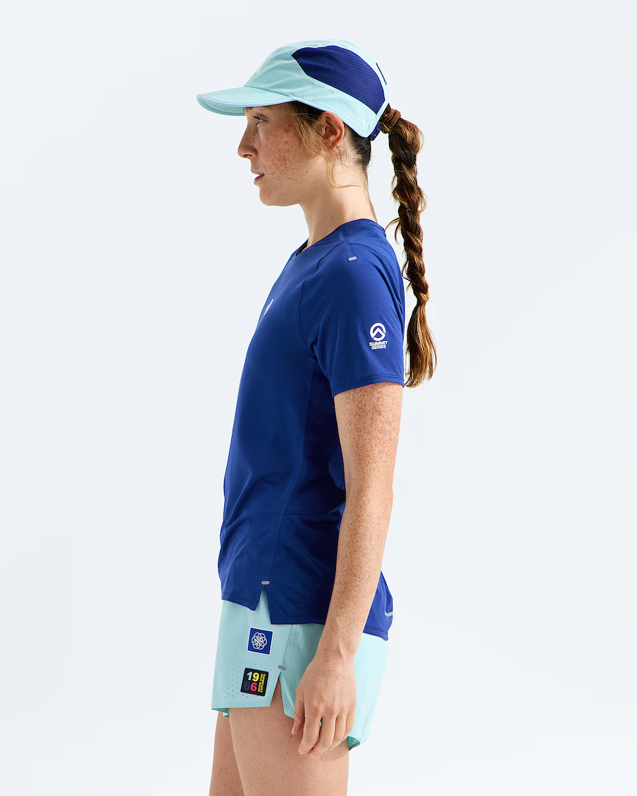 A woman in side profile wearing a royal blue athletic t-shirt, light blue athletic shorts, and a matching light blue visor against a clean white studio background.
