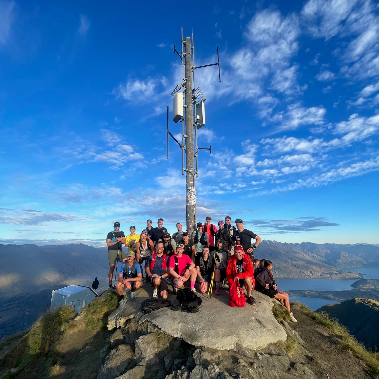 The Kinisi Froyday crew gathered at the summit of Roy's Peak under a bright blue sky, with Lake Wānaka below