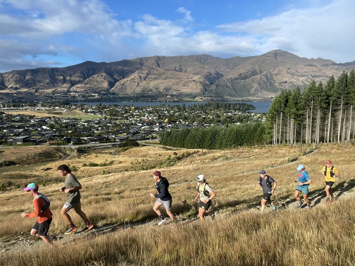 The Kinisi Running Sunday Long Run group running up a golden tussock trail above Wānaka, with the lake and mountains behind